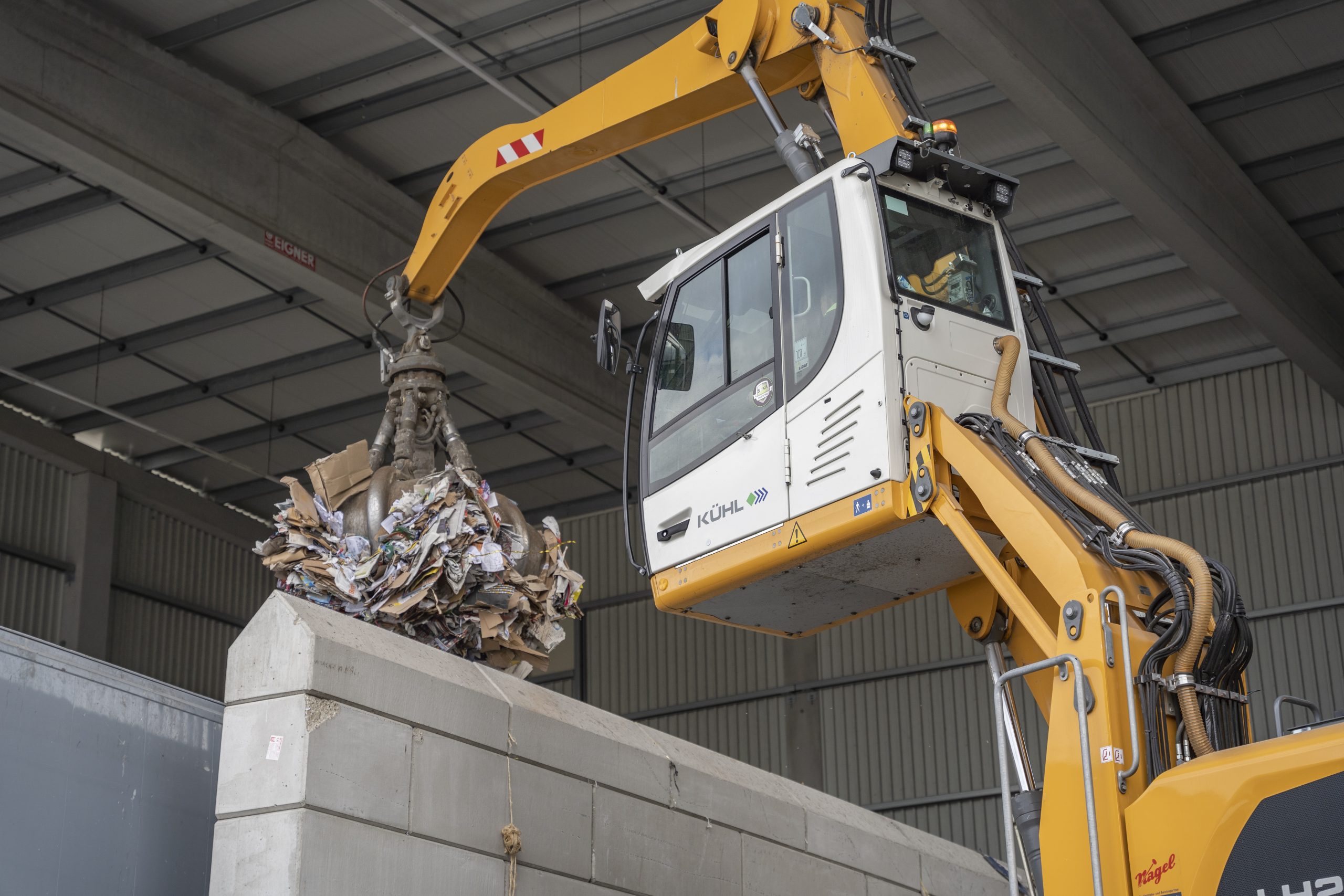 Bagger mit Greifer belädt einen Container mit Altpapier in der Halle